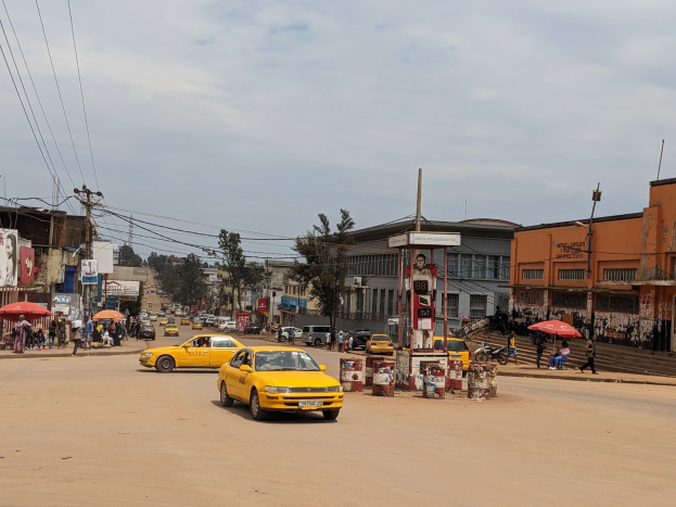 Ein gelbes Taxi fährt eine belebte Stadtstraße entlang, gesäumt von Gebäuden, Fußgängern mit Regenschirmen auf dem Gehweg und Strommasten mit Drähten im Hintergrund unter einem bewölkten Himmel.