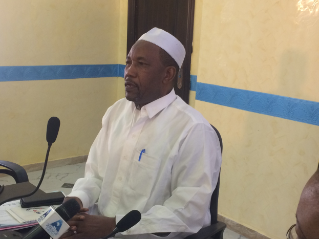 Ein Mann in einem weißen Kleid und Hut sitzt an einem Tisch mit Mikrofonen, spricht, während Papiere und Gegenstände auf dem Tisch liegen, und ein weiterer Mensch mit Brille rechts von ihm in einem Raum mit einer Wand und einer Tür im Hintergrund.