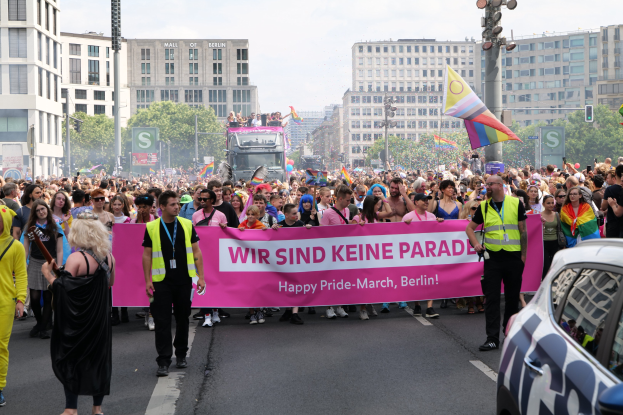 Eine große Gruppe von Menschen marschiert bei einer Pride-Veranstaltung in Berlin, die ein Banner mit der Aufschrift "Happy Pride March Berlin" trägt, mit einem geparkten Auto auf der rechten Seite und Gebäuden, Bäumen und einem klaren Himmel im Hintergrund.