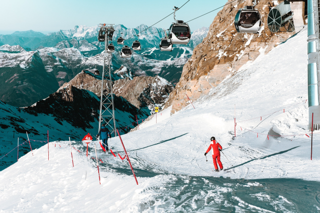 Gruppe von Skifahrern, die eine schneebedeckte Abfahrt mit einer Seilbahn und einer Gebirgskette im Hintergrund hinunterfahren, ein Skifahrer in roter Ausrüstung mit Skistöcken.