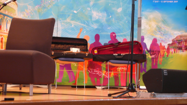 Auf einer Bühne stehen mehrere Stühle mit Gitarrenkoffern darauf, einige Mikrofone, ein Lautsprecher auf dem Boden und ein Banner im Hintergrund.