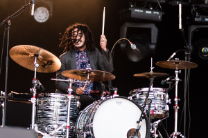 Ein Mann mit Dreadlocks, der auf der Bühne Schlagzeug spielt und Drumsticks mit Mikrofonständern vor sich hält, vor einem dunklen Hintergrund mit Bühnenlichtern.