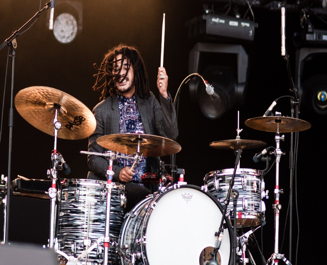 Ein Mann mit Dreadlocks, der auf der Bühne Schlagzeug spielt und Drumsticks mit Mikrofonständern vor sich hält, vor einem dunklen Hintergrund mit Bühnenlichtern.