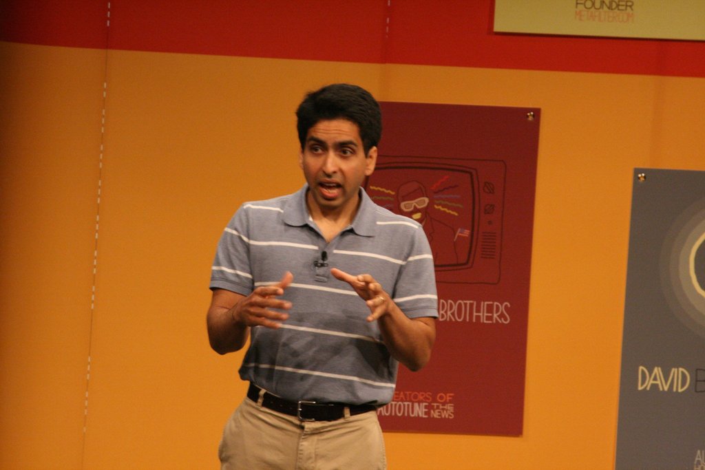 Ein Mann steht auf einer Bühne und spricht, mit einer Wand voller Plakate im Hintergrund.