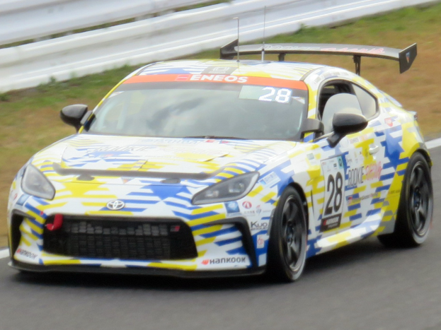 Ein weißer Toyota Supra GT3-Rennwagen auf einer Strecke mit Sponsor-Text, Rennnummern, einem Metallgeländer und Gras im Hintergrund.