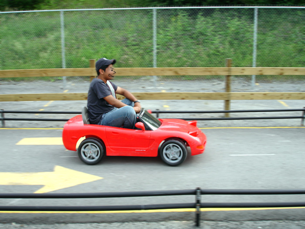 Ein Mann fährt ein kleines Auto auf einer Straße, mit Zaun und Bäumen im Hintergrund.