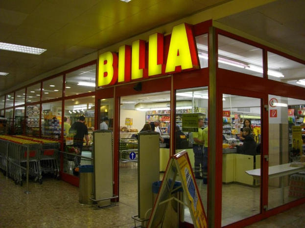 Ein Markt mit einem Eingang in der Mitte, Mülltonnen daneben, eine Reihe von Einkaufswagen auf der linken Seite, Deckenbeleuchtung und einige Menschen im Innern, die Produkte anschauen.