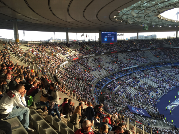 Große Menschenmenge in einem Stadion bei einem Fürballspiel mit einer Bühne, Fahnen, Masten und einem Bildschirm im Hintergrund.