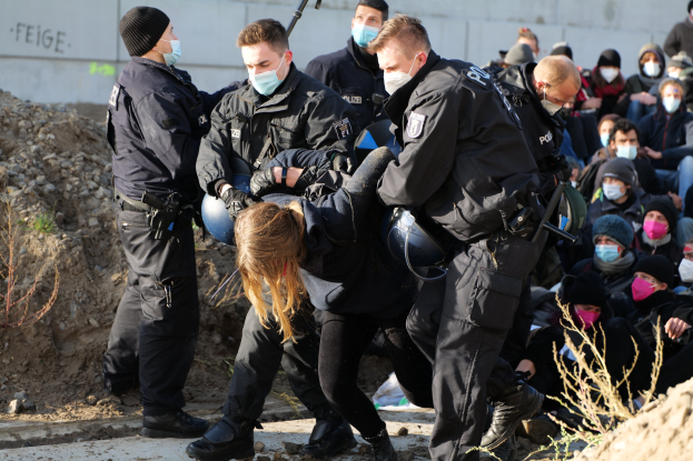 Gruppe maskierter Individuen in Käpfen, einige bewaffnet, umgeben eine Frau, die von der Polizei geführt wird, mit Pflanzen, Steinen und einer Wand mit Protesttext im Hintergrund.
