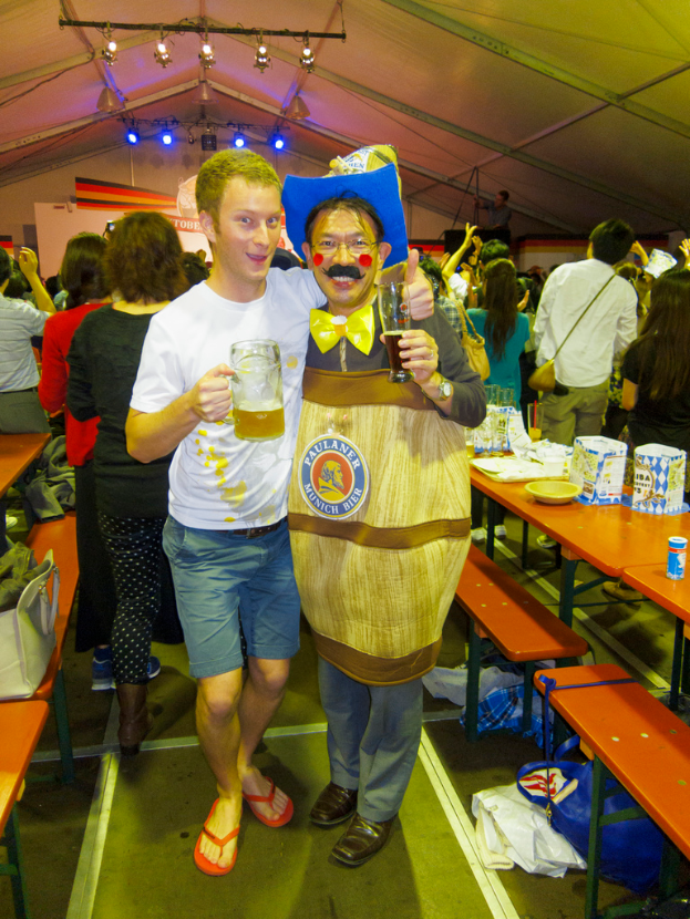 Zwei Personen stehen zusammen, eine hält ein Kind mit einem Bier, die andere trägt ein Theaterkostüm, daneben ein Tisch mit Essen und eine Gruppe von Menschen im Hintergrund.