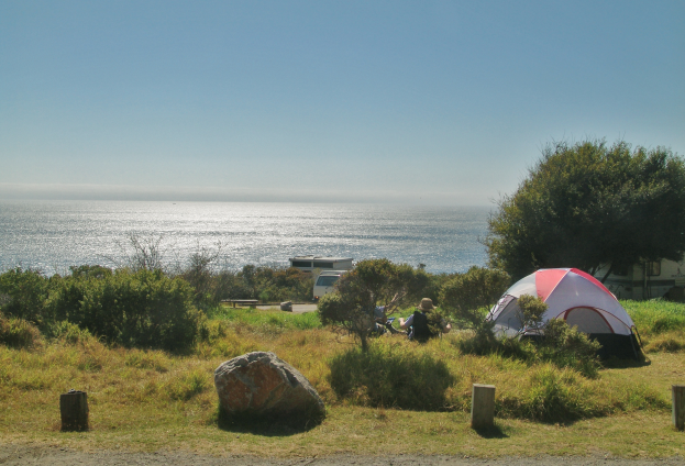 Ein Zeltplatz mit einem Zelt am Straßenrand neben dem Meer, mit ein paar Menschen um das Zelt sitzend, umgeben von Felsen, Pflanzen und Bäumen, mit einem Fahrzeug und der weiten Aussicht auf das Meer und den Himmel im Hintergrund.