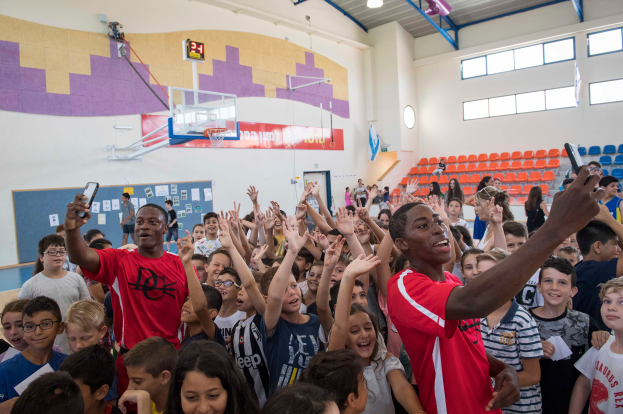 Eine Gruppe von Kindern steht vor einem Basketballfeld, einige halten Mobiltelefone, mit einer Tafel, Uhr, Torpfosten, Basketballkorb, Stühlen und Fenstern im Hintergrund.