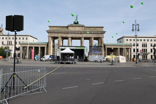 Brandenburger Tor in Berlin mit seinen Säulen und Statuen, umgeben von Gebäuden, Laternen, Verkehrszeichen, Zelten, Fahrzeugen, Menschen, Fahrrädern und grünen Luftballons vor einem bewölkten Himmel während des Berlin-Marathons.
