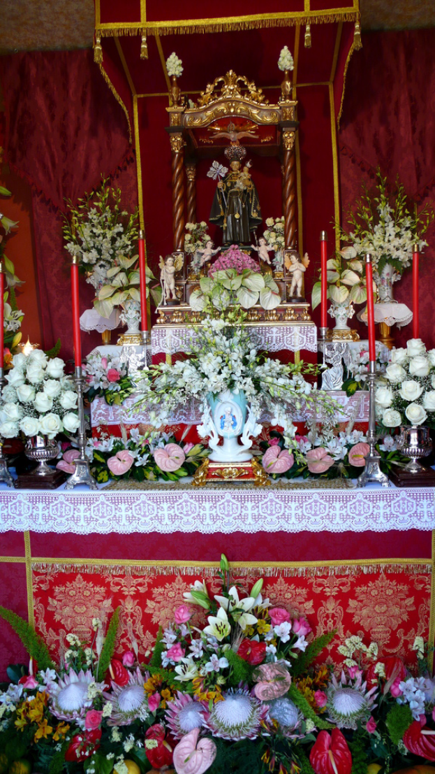 Idole auf einem Tisch mit Blumenvasen und Kerzenständern, die Kerzen halten, mit Blumen und Blättern unten und Vorhängen im Hintergrund.