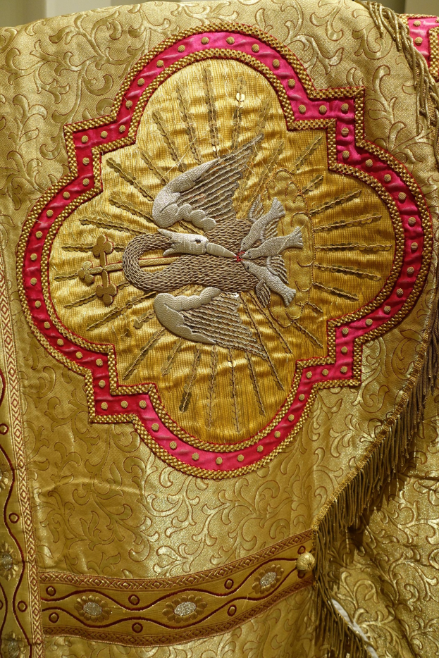 Nahaufnahme eines Priestergewandes mit aufwendiger Gold- und Rotstickerei und einem luxuriösen, königlich anmutenden Design vor einem neutralen Wandhintergrund.
