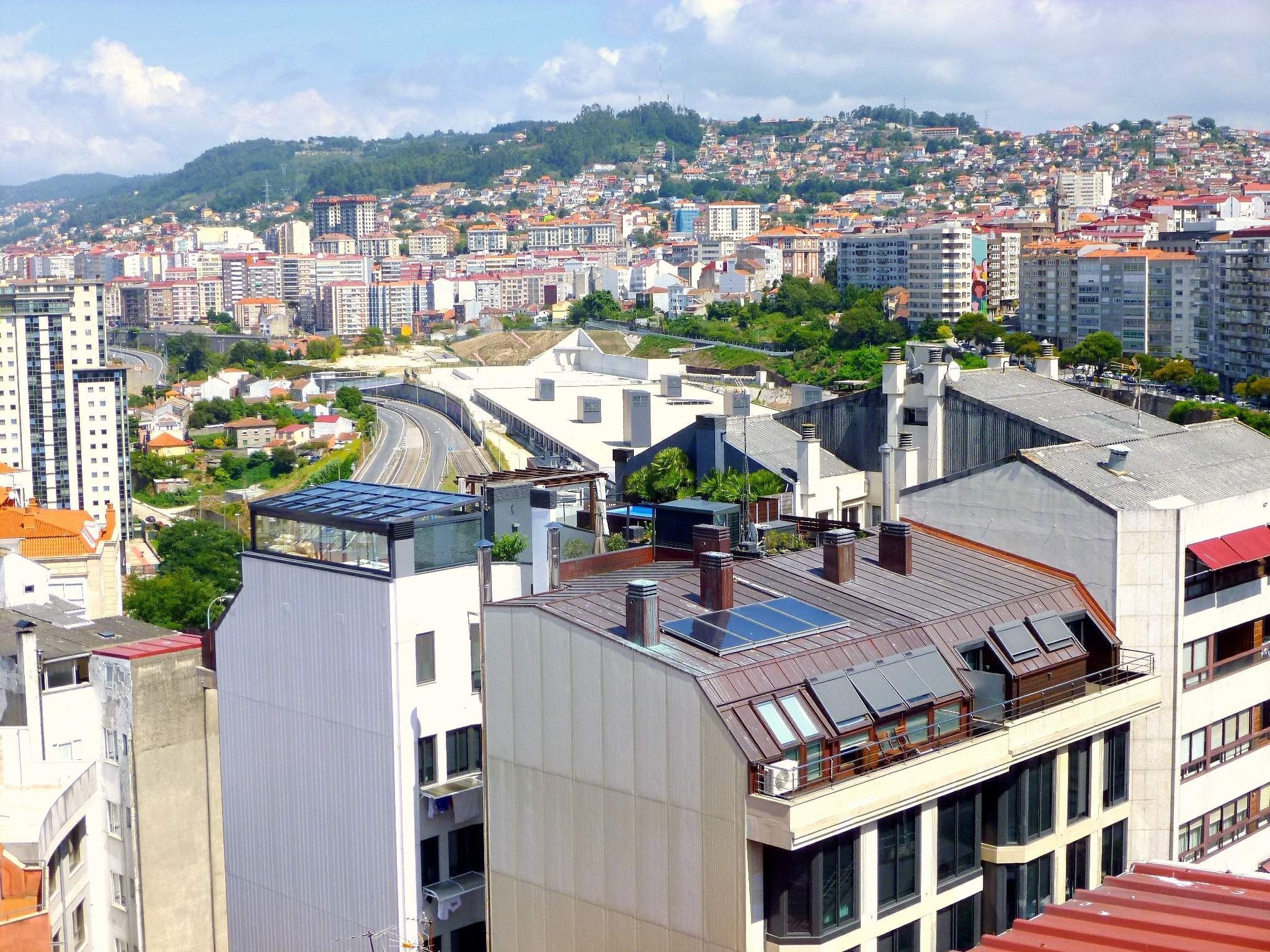 Luftaufnahme einer Stadt mit Gebäuden, Bäumen, Straßen, Fahrzeugen und Bergen, mit Solarpanelen auf einem Dach.