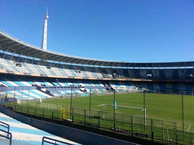 Großes Stadion mit einem von einem Zaun umgebenen Fußballfeld, ein Turm im Hintergrund und ein klarer blauer Himmel.