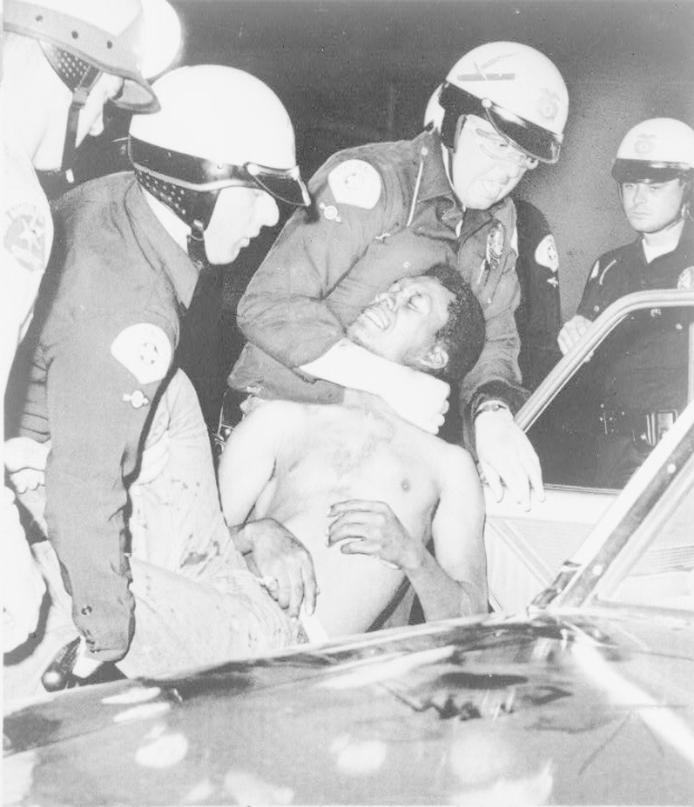 Ein Schwarz-Weiß-Foto eines besorgten Mannes, der von Polizisten in Helmen eskortiert wird, mit gesenktem Kopf und gefalteten Händen.