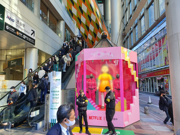 Eine Gruppe von Menschen steht vor einem Gebäude mit Glaswänden, Säulen und Texttafeln, einige tragen Masken, mit einer Rolltreppe im Blick; ein pinker Kasten in der Mitte zeigt Schaufensterpuppen und trägt den Text "Netflix's new Netflix Booth in Tokyo."