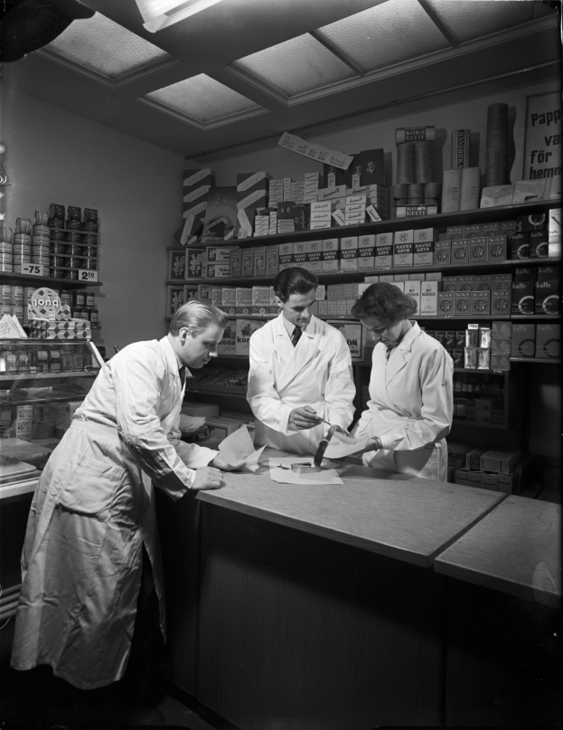 Drei Männer in weißen Kitteln stehen an einem Tresen in einer Apotheke mit Papieren und Gegenständen darauf, mit Regalen voller verschiedener Artikel im Hintergrund, in Schwarz-Weiß.