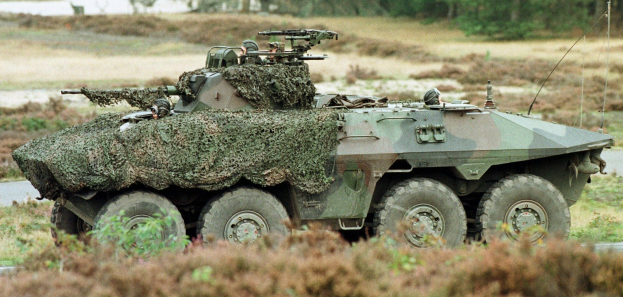 Militärpersonenwagen mit einer montierten großen Waffe, der auf einer Straße neben einem Wald fährt.