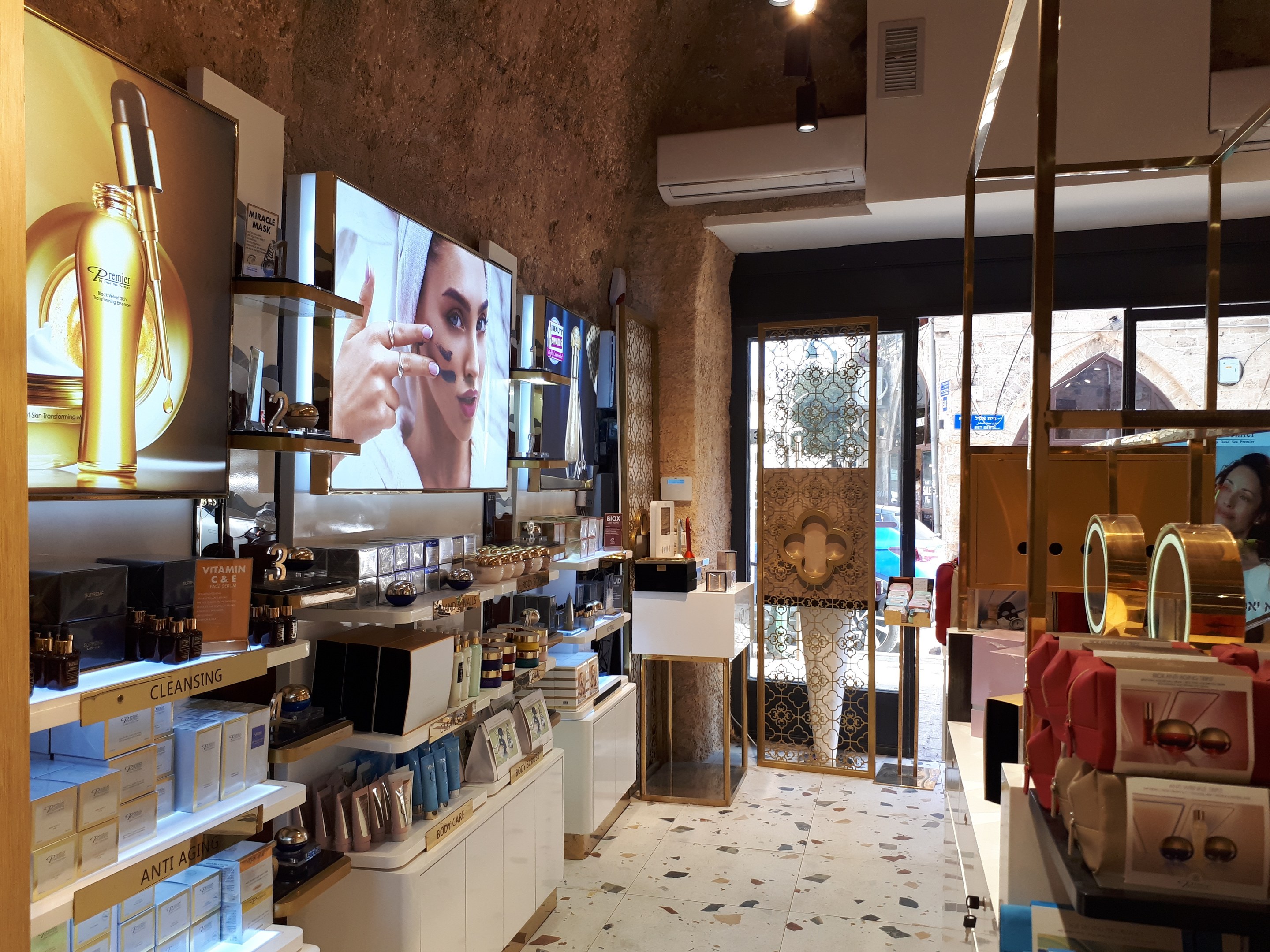 Ein gut beleuchteter Kosmetikgeschäftsinnenraum mit Regalen voller Produkte, an der Wand montierten Bildschirmen, Deckenleuchten, einer Klimaanlage und Glastüren, die eine Person, ein Auto und ein Gebäude draußen zeigen.
