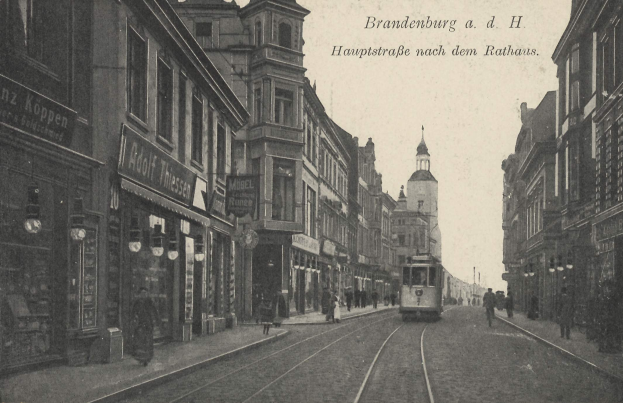 Ein Schwarz-Weiß-Foto einer Straßenkreuzung in Brandenburg, Deutschland, mit einer Straßenbahn auf den Schienen, Menschen auf dem Gehweg, Gebäuden mit Fenstern, Schildern und Straßenlaternen unter einem bewölkten Himmel.