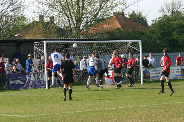 Spieler sind bei einem Fußballspiel auf einem Feld mit einem Tornetz beteiligt, während Zuschauer dahinter stehen, mit Bäumen und Häusern im Hintergrund.