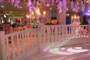 Eine Hochzeitsfeier mit einer weißen Balustrade, die mit Blumen geschmückt ist, Kronleuchter an der Decke, Menschen, die sich darum versammeln, Säulen und Lichter im Hintergrund und ein Bildschirm auf der rechten Seite.