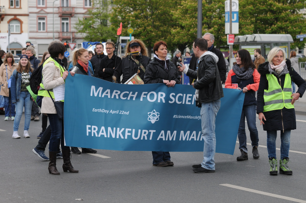 Gruppe von Menschen marschiert die Straße entlang und hält eine 'March for Science Frankfurt am Main'-Tafel mit Bäumen, Pfählen, Schildern, Gebäuden und einem klaren blauen Himmel im Hintergrund.
