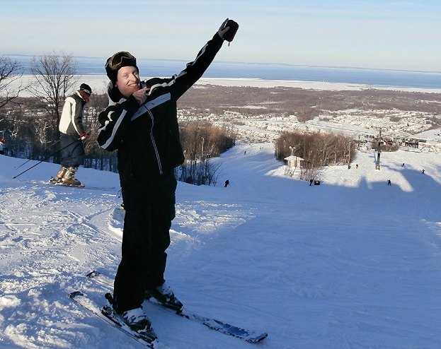 Eine Person auf einem Snowboard in der Mitte des Bildes, mit Skifahrern, Bäumen, Menschen, einem Pfahl, Wasser und Himmel im Hintergrund.