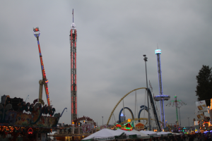 Eine Menschenmenge umringt einen hell erleuchteten Fasching bei Nacht, mit beleuchteten Fahrgeschäften, Pfählen und Bäumen unter einem bewölkten Himmel.