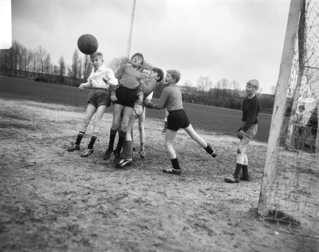 Schwarzes und weißes Foto von jungen Jungs, die auf einem Feld mit einem Torpfosten auf der rechten Seite und Bäumen im Hintergrund Fußball spielen.