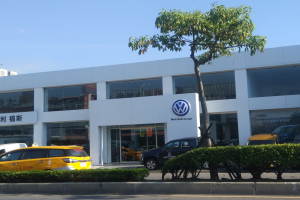 Ein VW-Autohaus mit parkenden Autos vorne, umgeben von Pflanzen und einem Baum, mit einem gläsernen Gebäude und einem sichtbaren Himmel im Hintergrund.