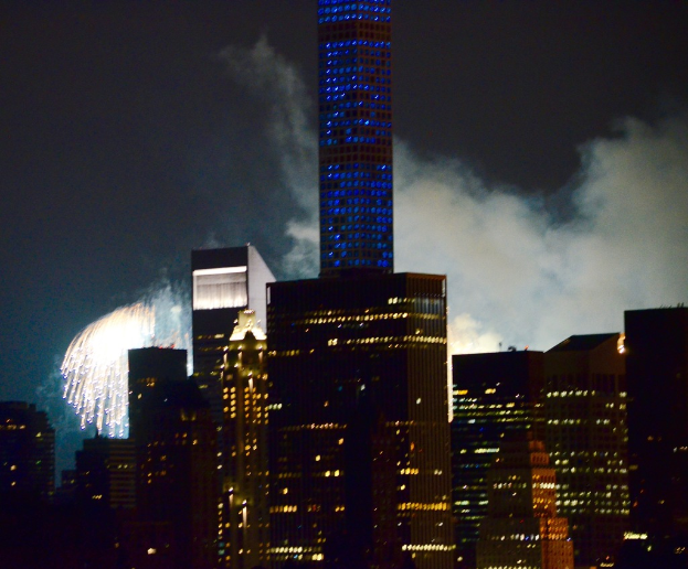 Gebäude mit Lichtern im Vordergrund, Feuerwerk und Rauch im Hintergrund, unter einem Himmel.