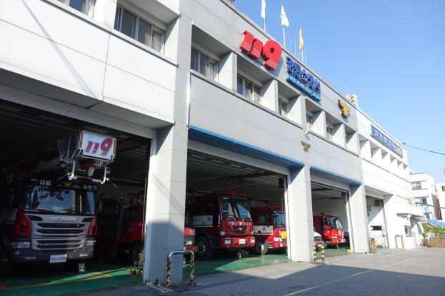 Feuerwache mit mehreren Feuerwehrautos vor dem Gebäude mit Fenstern, Säulen und Namensschildern, mit Flaggen im Vordergrund und anderen Gebäuden gegen einen klaren blauen Himmel.