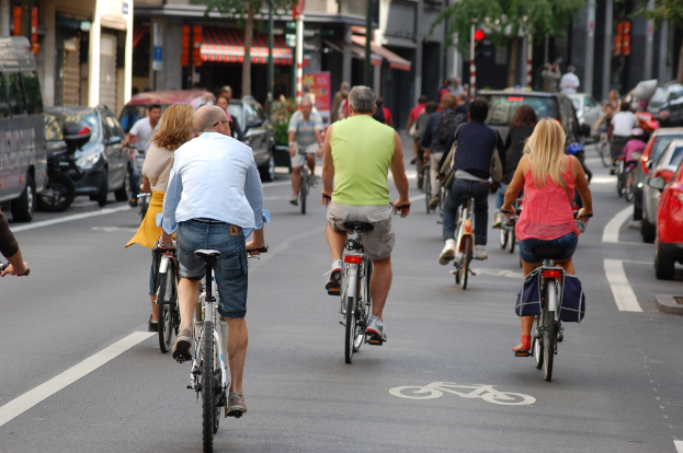 Menschen fahren Fahrrad auf Straßen mit Autos auf der linken und rechten Seite und Geschäften, einem Zelt, Bäumen und Gebäuden im Hintergrund.