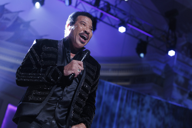 Lionel Richie performing at the Grammys, wearing a black dress and holding a microphone on a brightly lit stage.