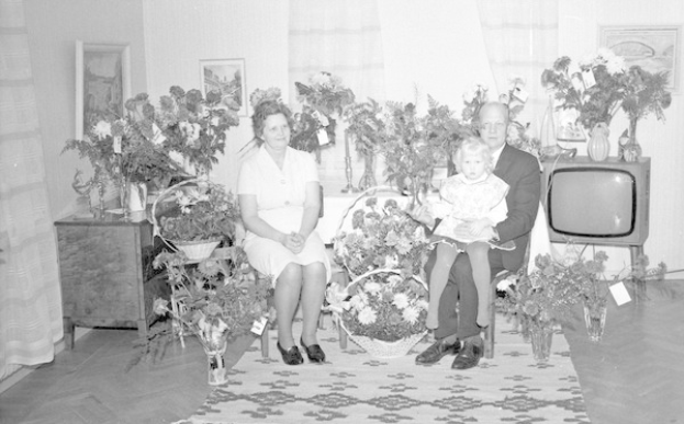 Schwarzes und weißes Foto einer Familie aus drei Personen - ein Mann hält ein Kind, eine Frau und das Kind sitzen auf einem Sofa mit Blumenvasen auf den Seitentischen, einem Teppich auf dem Boden, Vorhängen und gerahmten Fotos an der Wand.