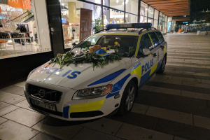 Ein mit Blumen und Blättern geschmückter Polizeiwagen parkt nachts vor einem Geschäft mit Glaswänden, das Schaufensterpuppen in Kleidern und beleuchtete Schilder zeigt.