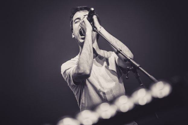 Ein Mann singt in ein Mikrofon auf einer hell erleuchteten Bühne, trägt ein schwarzes T-Shirt und Jeans mit stylischem, unordentlichem Haar, die Augen geschlossen in der Performance.