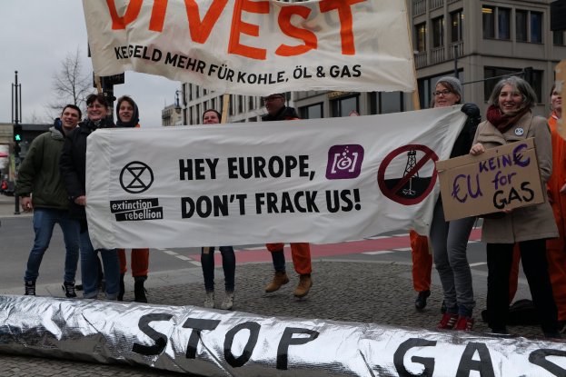 Eine Gruppe von Menschen steht auf einer Straße und hält ein Transparent mit der Aufschrift "Stoppt Gas" als Protest gegen die Europäische Union. Im Hintergrund sind Gebäude, Bäume, Pfosten, Verkehrsampeln und ein klarer blauer Himmel zu sehen.