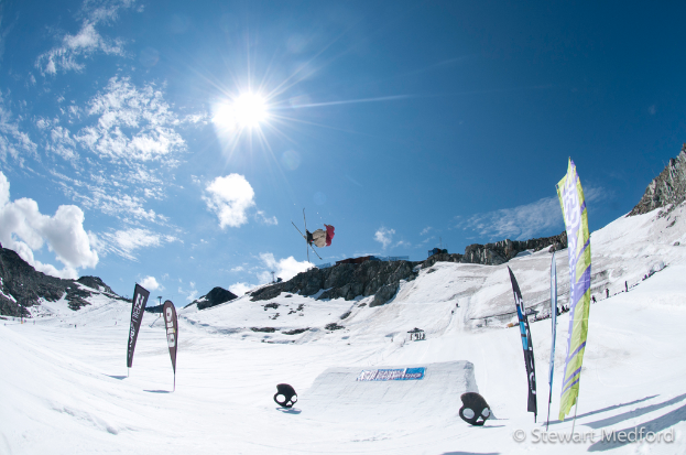 Eine Person in Skier steht in der Luft mit schneebedeckten Bergen im Hintergrund.
