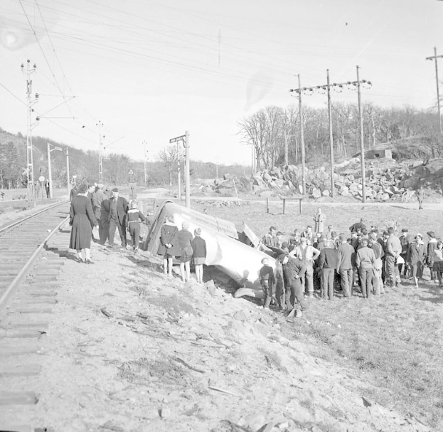 Eine Gruppe von Menschen steht um einen Zug, der auf einem Schotterhaufen auf einem Gleis gelandet ist, mit Strommasten und -leitungen im Hintergrund, Bäumen und Himmel in der Ferne, dargestellt in Schwarz-Weiß.