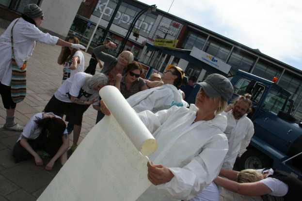 Eine Gruppe von Menschen, einige in Kostümen und Mützen, versammelt sich in der Mitte, wobei einige zu kämpfen scheinen, während eine Person ein Papier hält; im Hintergrund sind Gebäude, Pflanzen, Pfähle und Schilder mit Text, ein Fahrzeug auf der rechten Seite, ein Weg am Boden und der Himmel oben zu sehen.