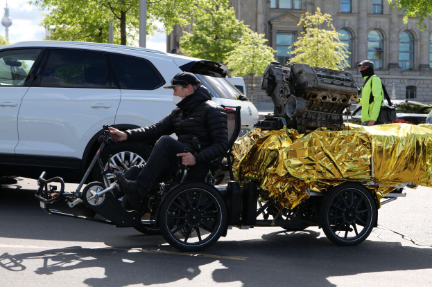 Ein Mann im Rollstuhl mit einem großen Motor auf dem Rücken, umgeben von Fahrzeugen auf einer Straße mit Bäumen, Gebäuden und einem klaren blauen Himmel im Hintergrund.
