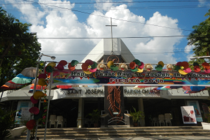 Ein Gebäude mit einer Tafel, die "Frohen Feiertag St. Joseph der Arbeiter" lautet, davor stehend, mit Treppen, einem bogenförmigen Eingang mit Dekorationen, Außenbestuhlung, Topfpflanzen, Bäumen und einem bewölkten Himmel.