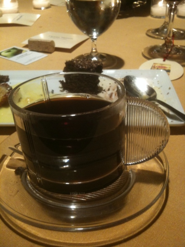 Ein Tisch mit Tasse, Untertasse, Teekanne, Teller, Löffel, Brownie, Glas, Wein, Kerze und weiteren Speisen.
