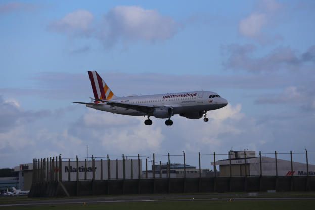 Germanwings Airbus A320-200 beim Starten am Frankfurter Flughafen, mit Gras darunter, einem Zaun im Vordergrund, Gebäuden und Bäumen im Hintergrund und einer bewölkten Himmel