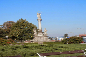 Ein Denkmal in einem Park mit zwei Bänken oben drauf, umgeben von Pflanzen, Gras, Bäumen, Straßenlaternen, einem Auto auf der Straße, Gebäuden und einem bewölkten Himmel.